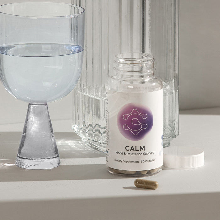 InfiniWell CALM mood support supplement bottle with open cap, capsule, and glass of water on tabletop.