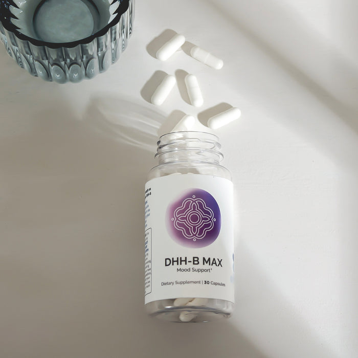 Infiniwell DHH-B MAX Mood Support supplement bottle with open cap, loose capsules and glass on light surface