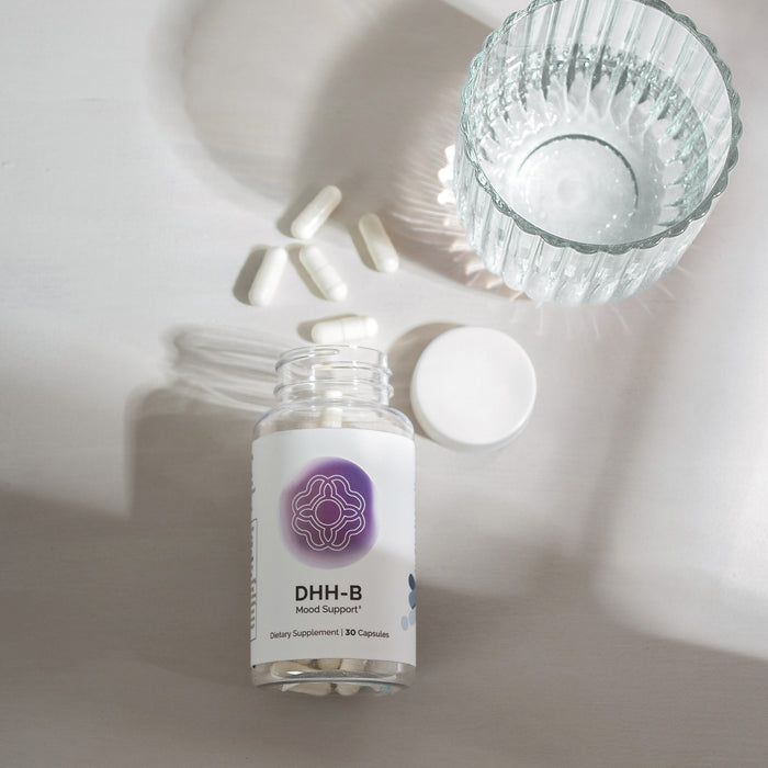 InfiniWell DHH-B Mood Support supplement bottle with open cap, white capsules, and textured glass of water on white surface.