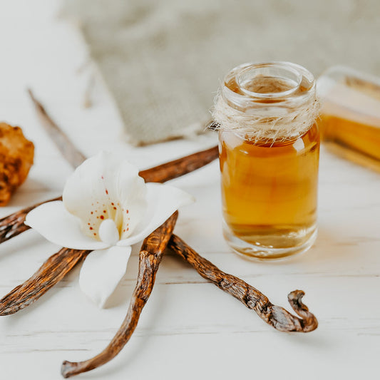 A small glass jar filled with golden vanilla extract, alongside vanilla beans and a white flower on a wooden surface.