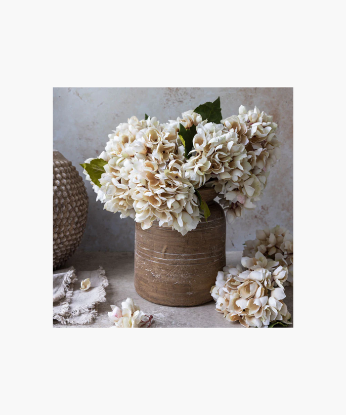 A bouquet of light pink hydrangea with lush blossoms and green leaves set in a terracotta pot, conveying a sense of delicate beauty. | SW Home