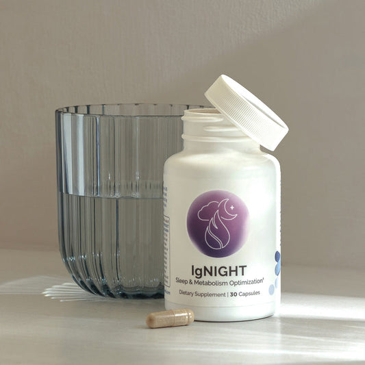Open bottle of InfiniWell IgNIGHT sleep and metabolism support supplement beside a glass with one capsule on the tabletop.