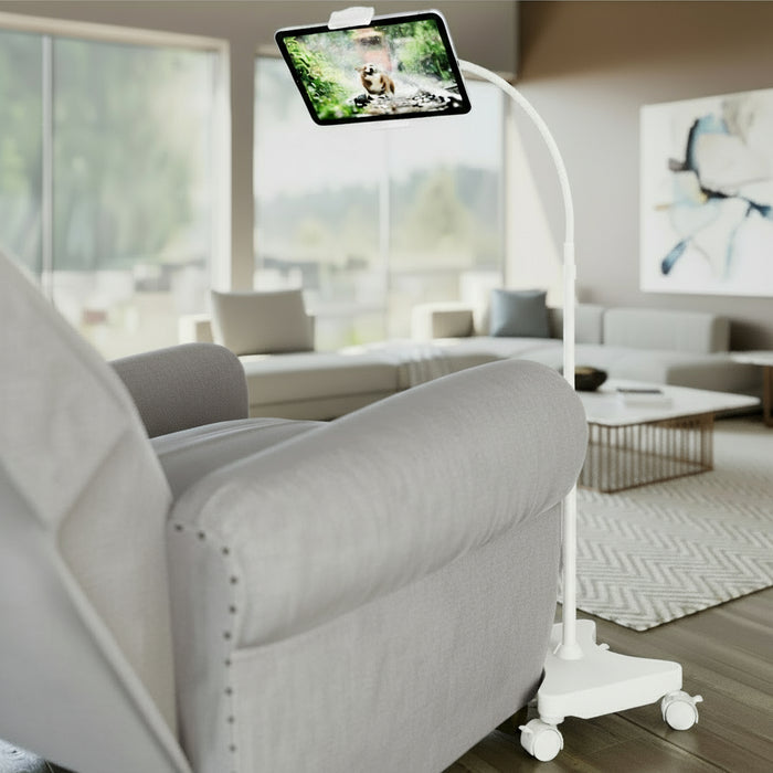 A black gooseneck tablet floor stand with casters, positioned next to a chair, displaying a tablet screen.