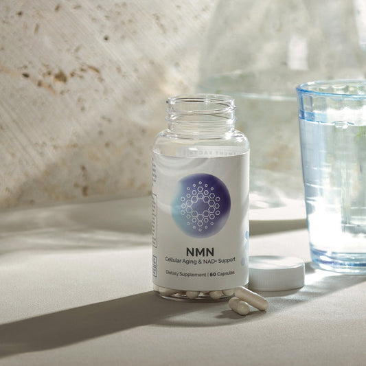 Infiniwell NMN cellular aging and NAD+ support supplement bottle with loose capsules, cap and glass of water nearby