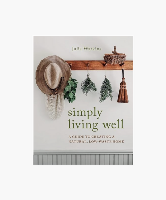 A book cover titled "Simply Living Well" by Julia Watkins, featuring a rustic wooden peg rail with a straw hat, herbs, basket, and broom hanging. The design conveys a natural, low-waste lifestyle theme. | Wildwood Home Co.