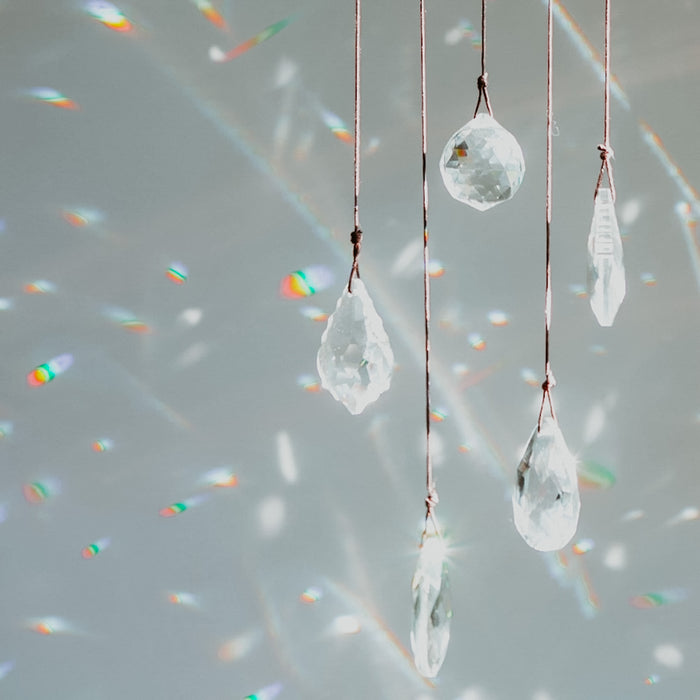 Transparent clear, teardrop-shaped crystals suspended on strings, with light reflecting off the bottles and creating rainbow-like patterns on a gray background.