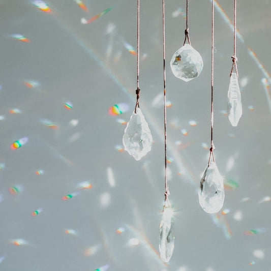 Transparent clear, teardrop-shaped crystals suspended on strings, with light reflecting off the bottles and creating rainbow-like patterns on a gray background.