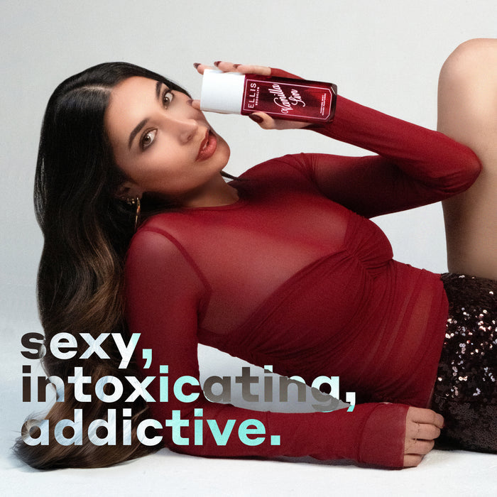 Model with brown hair wearing gold earrings, a red mesh shirt, and black sequined shorts lays down propped up on her arm while holding VANILLA SIN 240ml perfume mist on a white background with black, white, and blue text that reads "sexy, intoxicating, addictive."